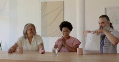 Çeşitli aileler masada hediye paketi açıyor, kız giysisini giyiyor, büyükanne de bebek için kart okuyor. Aile, kutlama, beklenti, sevinç, sıcaklık, samimiyet, birliktelik