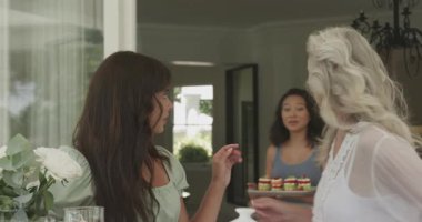 Farklı bayan arkadaşlar otururken veranda masasına çaydanlık yerleştirirken, toplanmak için tatlı koyarken. Kutlama, dostluk, sosyal toplantı, açık hava yemeği, çiçek süsü, şekerlemeler, sofistike