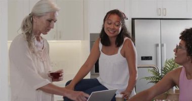 Dizüstü bilgisayarda el kol hareketleri yapan ve mutfak adasında çiçek taçı yapan çeşitli bayan arkadaşlar. El işi, işbirliği, diy, refah, hayat dolu, ev dekorasyonu, arkadaşlık.