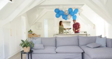 Bir çift adada şarap bardakları düzenliyor sonra da koltukta oturup balonların altında gülüyor. Romantik, kutlama, neşe, samimiyet, şenlik, boş zaman, ev