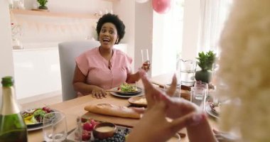 Çeşitli arkadaşlar öğle yemeğinin tadını çıkarıyorlar, yemek masasında, evde kahkaha ve sohbet paylaşıyorlar. Arkadaşlık, toplanma, sosyalleşme, yemek, eğlence, birliktelik.