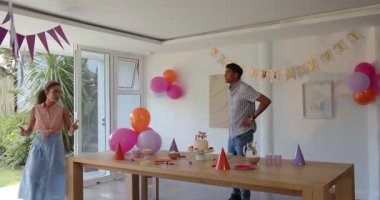 Çift, kapalı bir doğum günü partisinde kek, şapka ve atıştırmalıklarla dolu ahşap bir masanın yanında mutlu bir şekilde sohbet ediyor. Kutlama, balonlar, festival, birliktelik, neşe, parti, dekorasyon, nişan