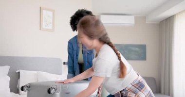 Afro-Amerikalı ve Asyalı arkadaşlar rahat yatak odasında gri bavullarını topluyor, seyahat kıyafetlerini sınıflandırıyorlar. Paketleme, arkadaşlar, yolculuk, konfor, yaşam tarzı, seyahat, organizasyon, hazırlık