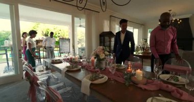 Farklı arkadaşlar verandanın cam kapılarına girip, akşam yemeği için uzun masanın etrafına akrilik sandalyeler diziyorlar. Zarif, kutlama, sofistike, sosyalleşme, mimari, dekor, ambiyans