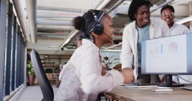 Afro-Amerikalı iş arkadaşları açık planlı ofiste kulaklık ve bar çizelgesiyle beşlik çakıyorlar. Kutlama, işbirliği, başarı, başarı, coşku, profesyonel, kurumsal