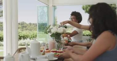 Güneş ışığı, veranda toplantısı için tatlı ve çay düzenleyen çeşitli bayan arkadaşlara kıvılcım saçıyor. Zarafet, dostluk, kutlama, misafirperverlik, çiçekli, barışçıl, şık