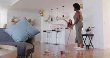 Afrikalı Amerikalı bir arkadaş mutfağı temizlerken Asyalı genç bir arkadaş parti dekorasyonu yapıyor. Temizlik, kutlama, arkadaşlar, mutfak, dekor, takım çalışması, hareketli, gündelik