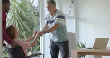 Üst düzey bir çift, erkek ona sandalyeye oturmasında yardım ederken ev sahibinin tekerlekli sandalyeyi itmesini selamlıyor. Bakıcı, arkadaşlık, bakım, ulaşılabilirlik, sağIık, modern, rahatlatıcı.