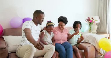 Parlak evdeki bej koltukta oturan Afro-Amerikan aile pastel balonlarla gülümsüyor. Kutlama, birliktelik, konfor, mutluluk, neşe, coşku, dekorasyon, bağ