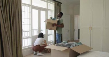 Asya kökenli Koreli ve Afrikalı Amerikalı çift yatak odasına kutularla giriyor, yerleşmek için yatak örtülerini açıyorlar. Taşınma, organizasyon, ev, dönüşüm, konfor, yaşam tarzı, iç mekan