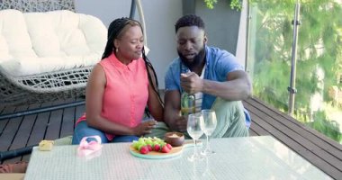 Afro-Amerikalı çift çilek seçiyor, çikolataya batırıyor, şarap dolduruyor, güvertede kadeh kaldırıyor. Romantik, boş zaman, zarafet, kutlama, mahremiyet, açık hava, yaşam tarzı
