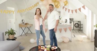 Yıldönümü pankartı gören, şarap içen, meyve besleyen ve evde dans eden son sınıf çifti. Romantizm, kutlama, yakınlık, neşe, şefkat, ev dekorasyonu, modern yaşam tarzı