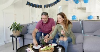 Çift, doğum gününü kanepede kutluyor. Meyve ve pasta tatmak için birbirlerini besleyip şarap yudumluyorlar. Romantik, kutlama, yakınlık, rahatlık, aile içi, birliktelik, neşe