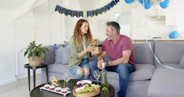 Kanepede oturan çift doğum günü afişini görüyor, hediye paketini değiştiriyor, açılıp kutluyor. Kutlama, samimiyet, neşe, eğlence, dekor, romantizm, mutluluk