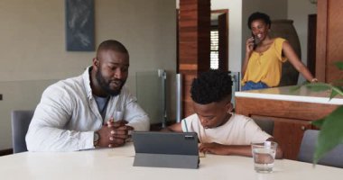 Baba oğluna tablet ödevinde yardım ediyor, anne telefonda konuşuyor. Aile, ebeveynlik, teknoloji, iletişim, çoklu görev, eğitim