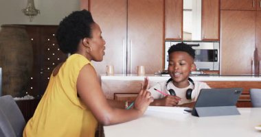 Anne oğluna tablet ödevinde yardım ederken, baba yakınlarda telefonla konuşuyor. Aile, ebeveynlik, teknoloji, eğitim, çoklu görev, yardım