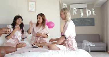 Farklı arkadaşlar gelini rahat yatak odasında pembe saten bornozlarla giydirip neşeli bir kutlama yapıyorlar. Kutlama, samimiyet, arkadaşlık, neşe, konfor, gelin, dekor, eşleştirme