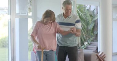 Üst düzey bir çift destek vermek ve bastonla yemek masasına doğru yürümek için kanepeden kalkıyor. Yaşlılar, bakım, arkadaşlık, hareket kabiliyeti, güven, destek, sıcaklık