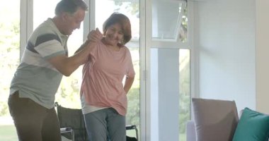 Su getiren Afrikalı Amerikalı hemşire yaşlı çifti tekerlekli sandalyeden kanepeye taşıdı. Bakıcı, destek, yardım, rehabilitasyon, şefkat, konfor, ev