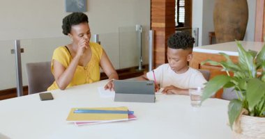 Anne, oğluna ödevlerinde yardım ediyor, tablet ve defterleri masada kullanıyor. Ebeveynlik, eğitim, öğrenim, teknoloji, aile