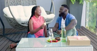 Balkondaki Afrikalı Amerikalı çift sarı kutuyu açıyor, sarılıyorlar ve büyük kurdeleli hediyeyi çözüyorlar. Romantik, kutlama, sevgi, sürpriz, neşe, açık hava, samimiyet