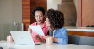 Evdeki belgeleri inceliyor, Afro-Amerikan kadınları dizüstü bilgisayar kullanıyor ve birlikte gülümsüyor. Uzaktan çalışma, işbirliği, teknoloji, ev ofisi, çeşitlilik, çoklu görev