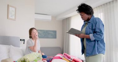 Afro-Amerikalı ve Asyalı arkadaşlar yatak odasında kıyafet paketliyor, seyahat sırasında cep telefonu kullanıyorlar. Paketleme, arkadaşlık, seyahat, giysi, sohbet, yatak odası, planlama, canlı