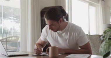 Adam belgeleri inceliyor, notları karalıyor ve evdeki masada kahve fincanını yudumlarken dizüstü bilgisayarda yazıyor. Minimalist, üretkenlik, çalışma alanı, konsantrasyon, yalnızlık, doğal, samimi