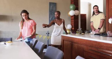 Arkadaşlar partiye hazırlanıyor, sofra kuruyor ve neşeli ifadelerle yemek servisi yapıyorlar. Kutlama, toplanma, mutluluk, yemek, şenlik, arkadaşlık