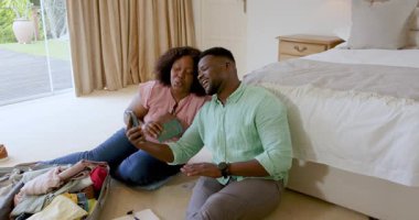 Yatak odasında oturmuş bavullarını toplarken selfie çeken gülümseyen bir çift. Seyahat, tatil, planlama, teknoloji, mutluluk