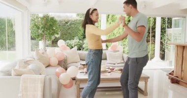 Genç çift oturma odasında balonlarla ve pastayla neşeyle dans ediyor. Kutlama, parti, birliktelik, mutluluk, keyif, aşk