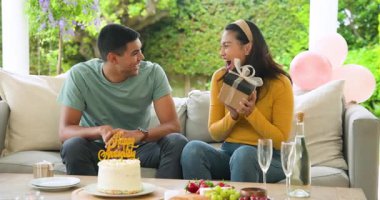 Kanepede hediyeleşen genç bir çift, doğum gününü pasta ve şampanyayla kutluyorlar. Kutlama, romantizm, yıldönümü, sürpriz, beraberlik, hediye