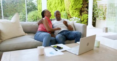 Siyahi adam ve kadın konuşuyor, kağıtları kontrol ediyor, gülümsüyor, kucaklıyor ve evdeki kanepede oturan dizüstü bilgisayarını kullanıyor. Ev hayatı, ilişki ve yaşam tarzı kavramı.