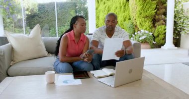 Siyahi adam ve kadın konuşuyor, kağıtları kontrol ediyor, hesap makinesi ve dizüstü bilgisayarla evdeki kanepede oturuyor. Ev hayatı, ilişki ve yaşam tarzı kavramı.