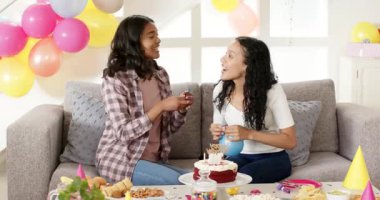 Farklı kız kardeşler oturma odasındaki kanepede mavi balonlar şişiriyorlar, doğum günü pastası ve atıştırmalıklarıyla. Kutlama, şenlik, pastel, canlı, bağlanma, dekorasyon, ev