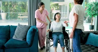 Tekerlekli sandalyede oturan mutlu son sınıf öğrencisi kadın, kadın hemşire ve arkadaşıyla oturma odasında konuşuyor. Kıdemli yaşam tarzı, sakatlık, sağlık ve arkadaşlık konsepti.