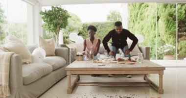 Afro-Amerikalı çift gülüyor ve oturma odasında kutlama masası kuruyor. Toplantı, yemek, mutluluk, eğlence, neşe, dekor