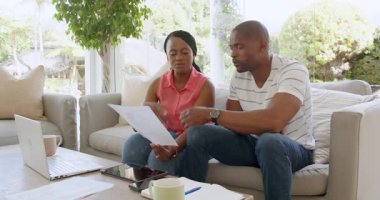 Siyahi adam ve kadın konuşuyor, kağıtları kontrol ediyor, akıllı telefon ve dizüstü bilgisayarla evdeki kanepede oturuyor. Ev hayatı, ilişki ve yaşam tarzı kavramı.