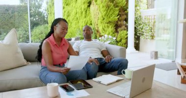Siyahi adam ve kadın konuşuyor, kağıtları kontrol ediyor, hesap makinesi ve dizüstü bilgisayarla evdeki kanepede oturuyor. Ev hayatı, ilişki ve yaşam tarzı kavramı.
