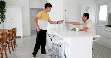 Yaşlı adam mutfak tezgahından izlerken genç adam kanepede zarf açıyor. Ev, aile, oturma odası, iletişim, ebeveynlik, etkileşim