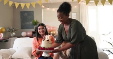 Üç mutlu çok ırklı ve siyah bayan arkadaş güneşli oturma odasındaki bir doğum günü partisinde pastaya mum üflüyorlar. Kutlama, doğum günü, parti, yaz ve arkadaşlık kavramı.