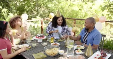 Bir grup mutlu, çok ırklı, siyah ve Çinli erkek ve kadın arkadaş terasta bir doğum günü partisinde şampanya açıyorlar. Kutlama, doğum günü, parti, yaz ve arkadaşlık kavramı.