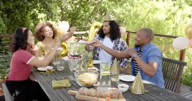 Bir grup mutlu, çok ırklı, siyah ve Çinli erkek ve kadın arkadaş masada oturup terastaki bir doğum günü partisinde yemek yiyorlar. Kutlama, doğum günü, parti, yaz ve arkadaşlık kavramı.