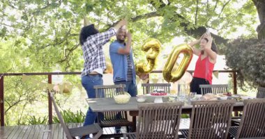 Bir grup mutlu, çok ırklı, siyah ve Çinli erkek ve kadın arkadaş terasta bir doğum günü partisi için sofra ve dekorasyon hazırlıyorlar. Kutlama, doğum günü, parti, yaz ve arkadaşlık kavramı.