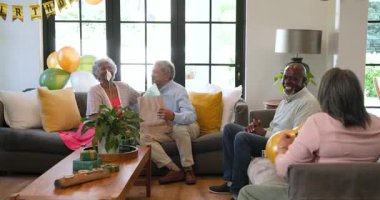 Bir grup mutlu üst düzey çok ırklı, beyaz ve siyah erkek ve kadın arkadaş oturma odasındaki bir doğum günü partisinde hediye veriyorlar. Kutlama, doğum günü, parti, son sınıf yaşam tarzı ve arkadaşlık konsepti.