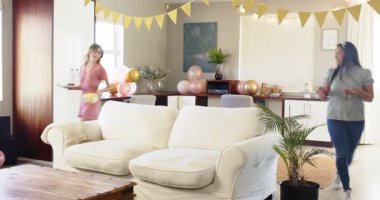 İki mutlu çok ırklı ve beyaz bayan arkadaş güneşli oturma odasında bir doğum günü partisi için masa ve dekorasyon hazırlıyor. Kutlama, doğum günü, parti, yaz ve arkadaşlık kavramı.