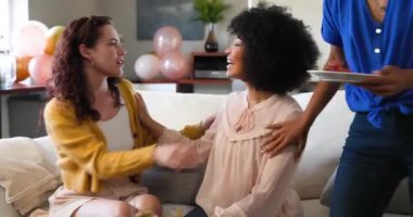 Üç mutlu çok ırklı ve beyaz bayan arkadaş güneşli oturma odasında bir doğum günü partisinde konuşup kucaklaşıyorlar. Kutlama, doğum günü, parti, yaz ve arkadaşlık kavramı.