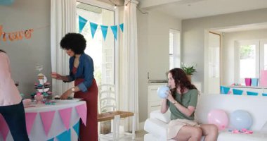 Üç mutlu çok ırklı ve beyaz bayan arkadaş balonları şişiriyor ve güneşli oturma odasında bebek partisi hazırlıyor. Kutlama, annelik, parti, yaz ve arkadaşlık kavramı.