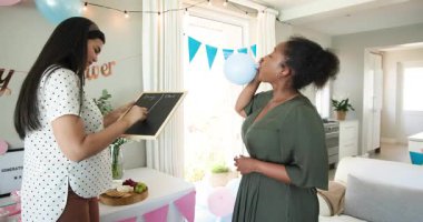 İki mutlu çok ırklı ve siyah bayan arkadaş balonları şişirip güneşli oturma odasında bebek partisine hazırlanıyor. Kutlama, annelik, parti, yaz ve arkadaşlık kavramı.