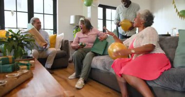 Bir grup mutlu üst düzey çok ırklı, beyaz ve siyah erkek ve kadın arkadaş oturma odasındaki bir doğum günü partisinde hediye veriyorlar. Kutlama, doğum günü, parti, son sınıf yaşam tarzı ve arkadaşlık konsepti.